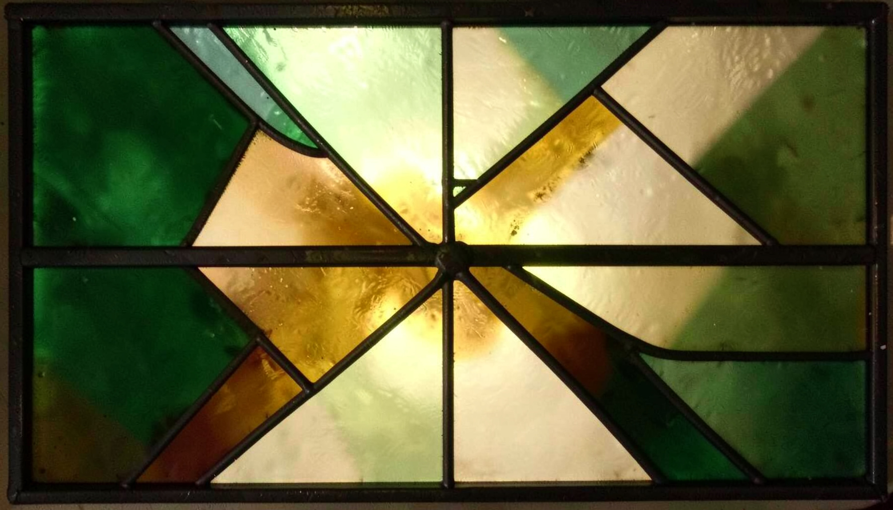 A stained-glass composition with bold leaded segments in deep greens, amber golds, and muted ivory, forming an abstract geometric pattern.